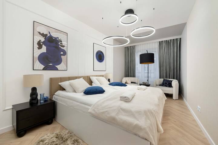 Spavaća soba s velikim krevetom, plavo-bijelom posteljinom i apstraktnim zidnim umjetničkim djelima. Prozor s zavjesama pruža mekanu rasvjetu.