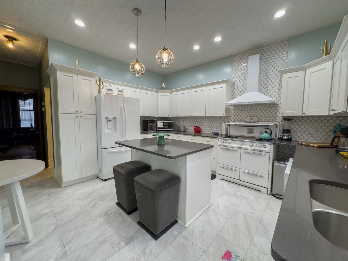 A modern kitchen features white cabinetry and a large island with two dark stools. Stainless steel appliances are complemented by a patterned backsplash. Ample overhead lighting illuminates the space, and a light blue paint adds a soft touch to the ambiance.