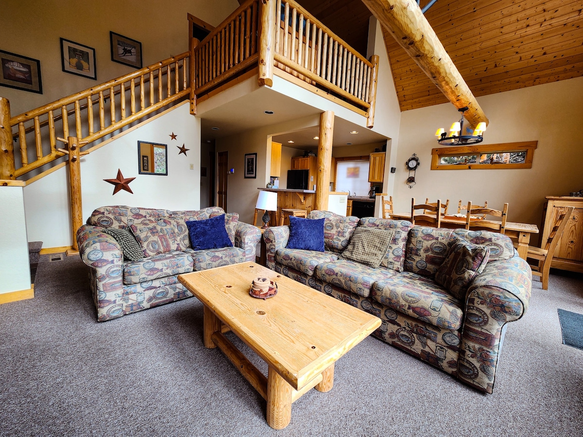 An open living space is highlighted by high ceilings and exposed wooden beams. Two patterned sofas, adorned with accent pillows, face a rustic wooden coffee table. The area features a cozy atmosphere, with artwork on the walls and a view into the kitchen and dining area.