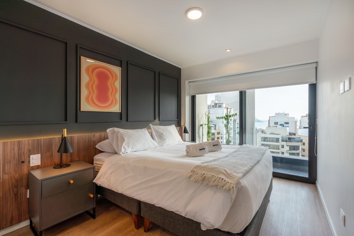 A spacious bedroom features a large bed with crisp white linens and a light-colored throw draped over the side. A nightstand with a lamp stands beside the bed. Floor-to-ceiling windows allow natural light to fill the room and provide a view of the surrounding city.
