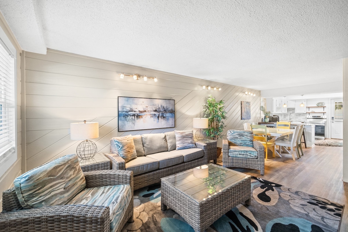A bright living area features woven-style seating with soft cushions, providing a comfortable environment. A coffee table sits atop a patterned rug, with light streaming in through a window. The open layout reveals a dining area and kitchen in the background.