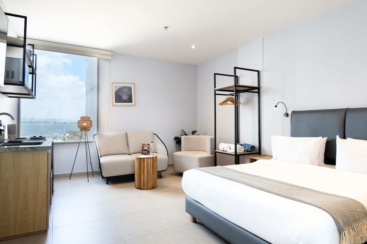 A modern studio apartment is filled with natural light from a large window showcasing views of the city. The space includes a comfortable bed with quality linens, a cozy seating area with a sofa, and a small wooden table. A functional kitchenette is visible, equipped with essential appliances.