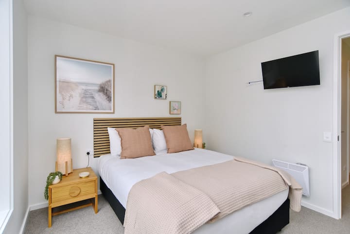 Chumba cha kulala chenye starehe na utulivu chenye kitanda cha kifahari, rangi laini zisizoegemea upande wowote, mapambo maridadi na televisheni iliyowekwa ukutani kwa ajili ya burudani na mapumziko yako