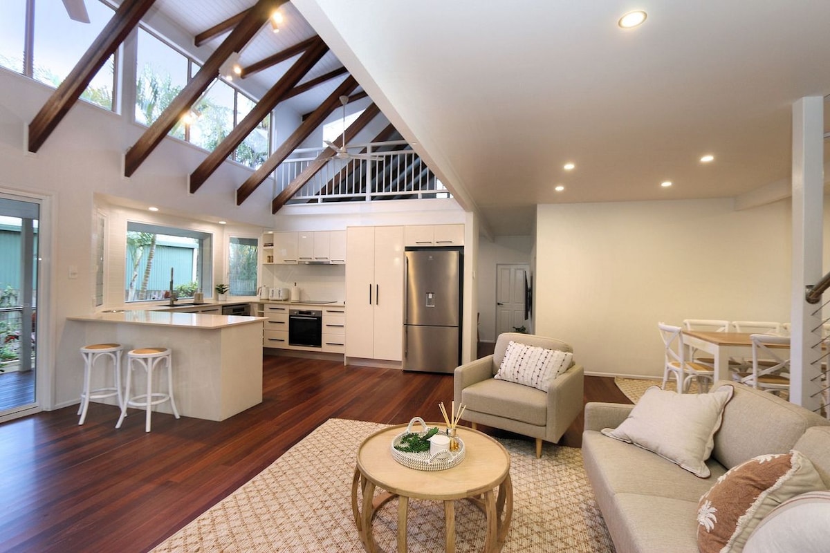 An open-concept living area features high ceilings with exposed beams and warm wooden floors. A cozy seating arrangement surrounds a circular coffee table, while a modern kitchen with sleek cabinetry and stainless-steel appliances is visible in the background. Natural light fills the space from large windows.