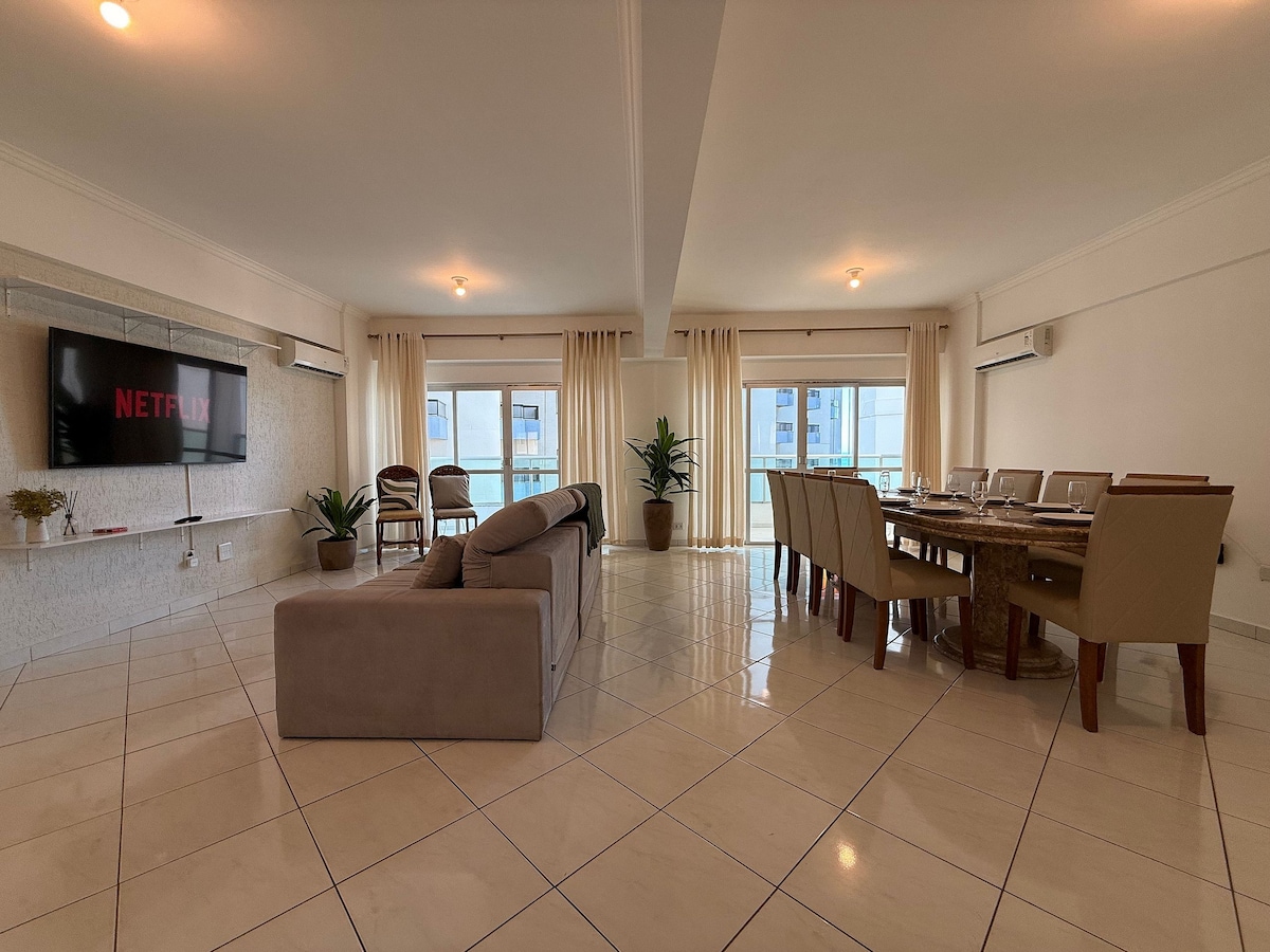 A spacious living area features a large sofa and a dining table for ten. Floor-to-ceiling windows allow natural light to fill the room, complemented by light-colored curtains. A smart TV is mounted on the wall, providing easy access to various streaming services.