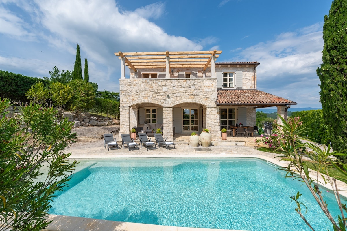 The two-story villa is surrounded by lush greenery and features a private swimming pool with a sun terrace. Sun loungers are arranged next to the pool, while the upper balcony offers an outdoor seating area under a wooden pergola. The natural stone façade adds to the charm.