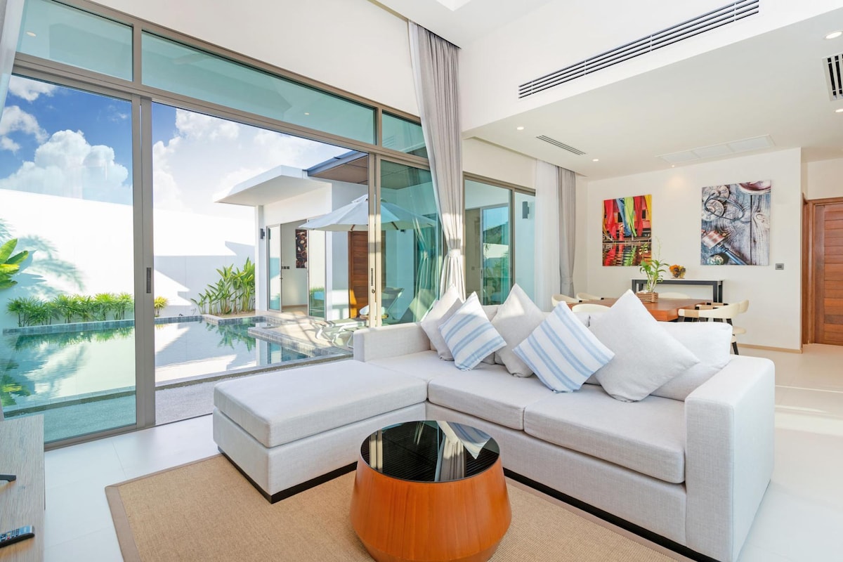 A modern living area features a spacious sectional sofa with light-colored cushions, paired with a round wooden coffee table. Large glass doors provide a view of the private pool, enhancing the indoor-outdoor connection. Bright artwork adorns the walls, contributing to the room's contemporary design.