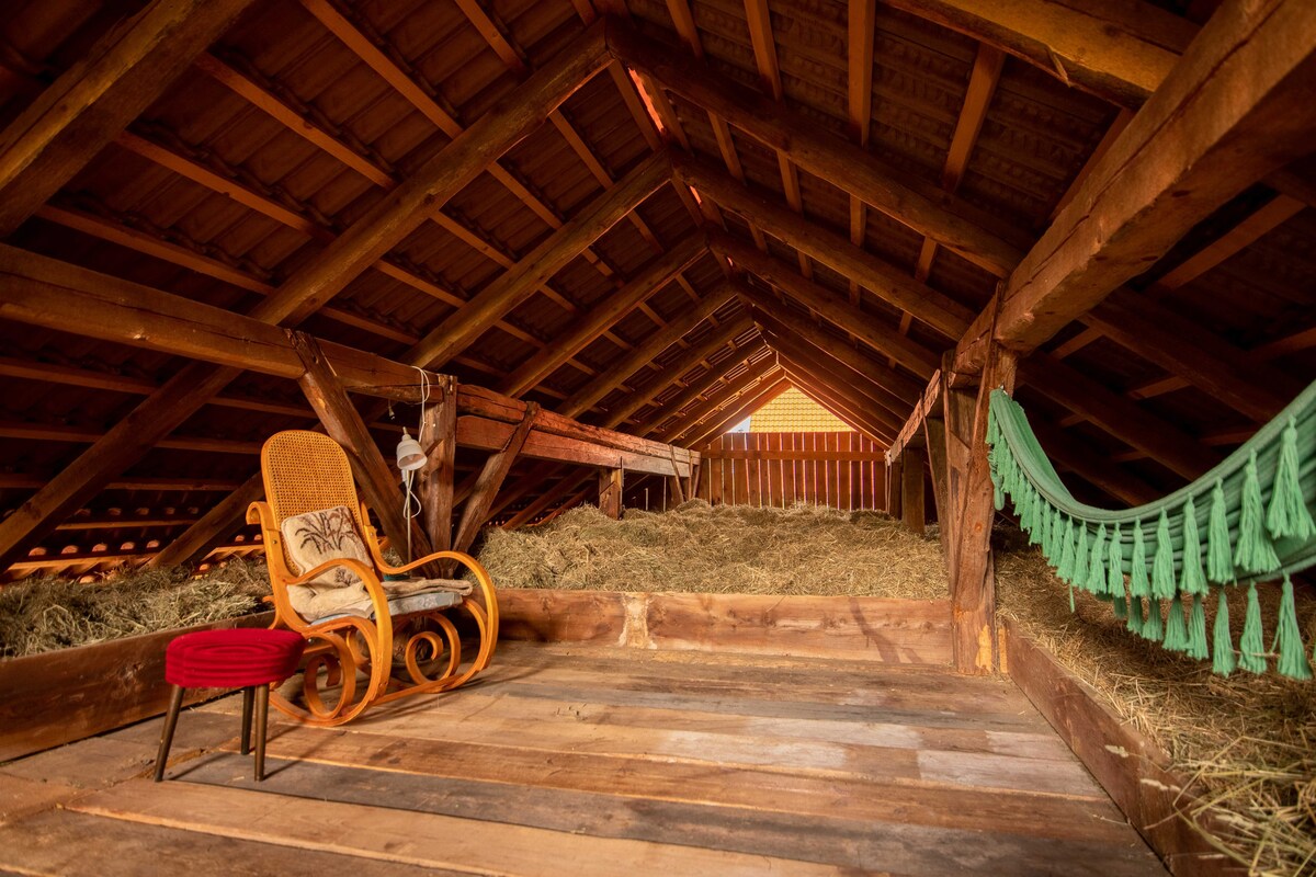 The attic space features exposed wooden beams and a rustic wooden floor, filled with fragrant hay. A single decorative chair is positioned against one wall, while a green textile swag hangs decoratively. The area offers a serene and cozy atmosphere for relaxation.