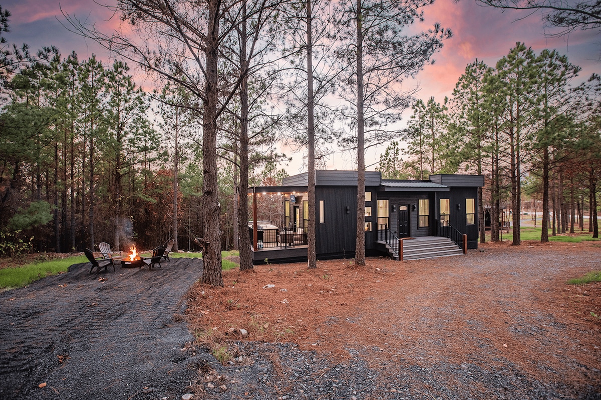 A modern cabin is nestled among tall pine trees, featuring a dark exterior and large windows that invite natural light. A gravel pathway leads to a spacious deck, while a firepit area with seating is visible nearby, creating an inviting outdoor space.