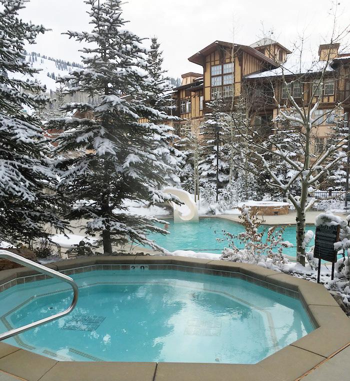 A hot tub is shown in the foreground, surrounded by snow-covered trees. In the background, a pool is visible, creating a serene winter scene. The architectural details of the nearby building feature tall windows and a rustic aesthetic, enhancing the tranquil atmosphere.