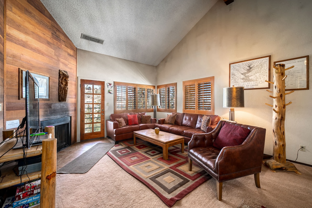 A spacious living room features a high vaulted ceiling with natural light coming through multiple windows. A large sofa and armchairs are arranged around a wooden coffee table, with a warm-colored area rug underneath. A flat-screen TV is situated on a side unit with a wood burning fireplace nearby.