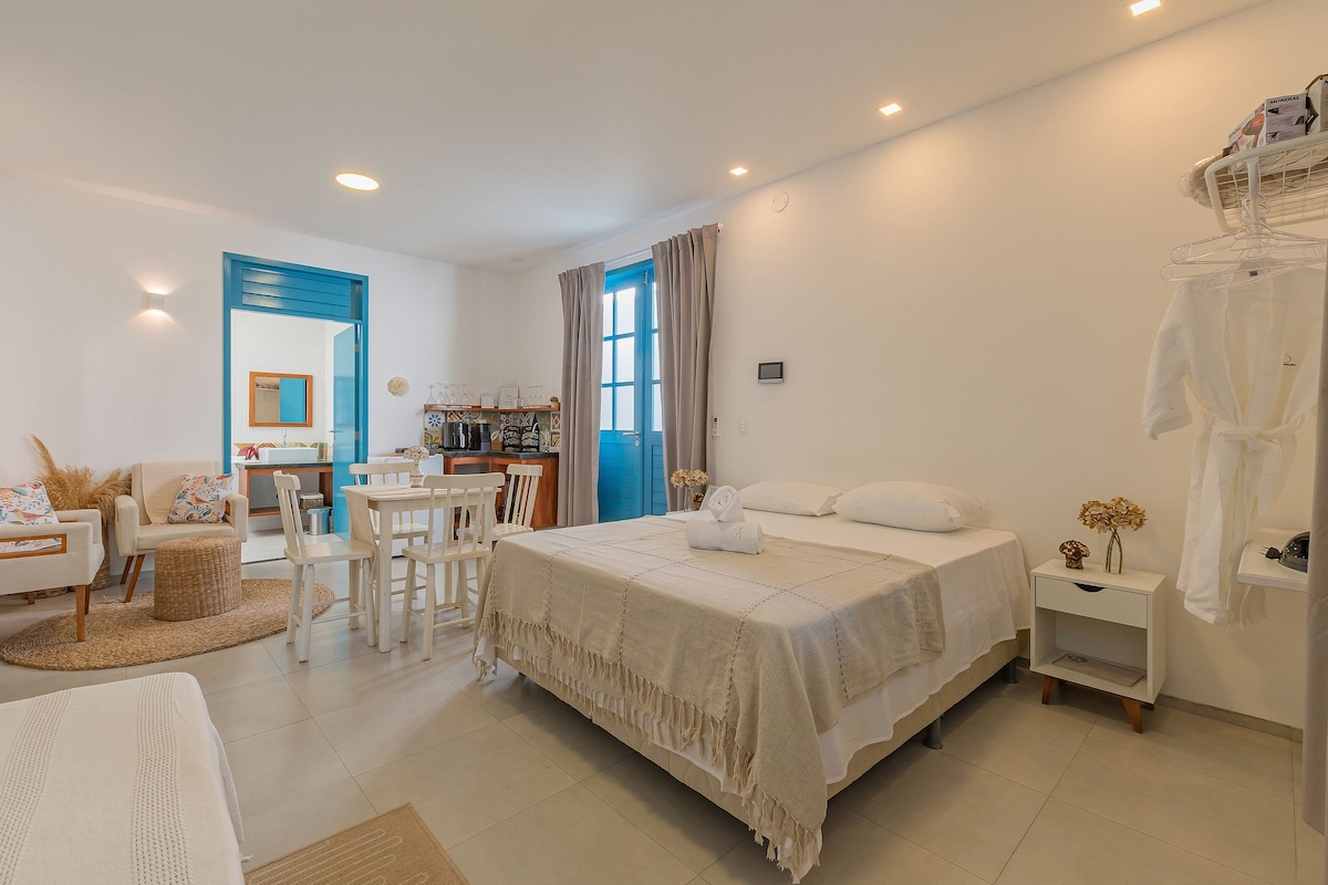 A bright and airy suite features a spacious bed with neutral bedding, complemented by a light-coloured area rug. A dining table and chairs are positioned nearby, with a cozy seating area in the corner. Large windows allow natural light to fill the room.