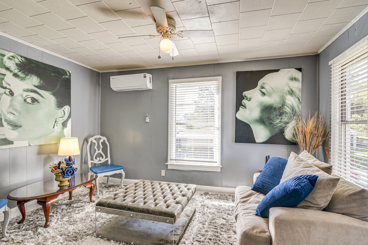 A comfortable living area is presented with a plush, tufted coffee table at the center and a light-colored sofa adorned with blue cushions. Windows allow natural light to brighten the space, complemented by a ceiling fan. Decor features modern wall art and decorative plants.