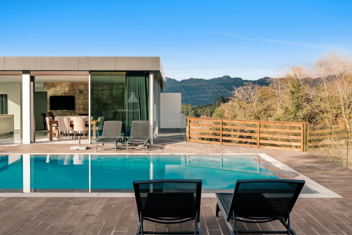 The outdoor area showcases a private pool with clear water, flanked by two lounge chairs. A modern structure with large glass panels reflects natural light, and distant mountains provide a scenic backdrop. Wooden fencing delineates the space, enhancing the tranquil atmosphere.