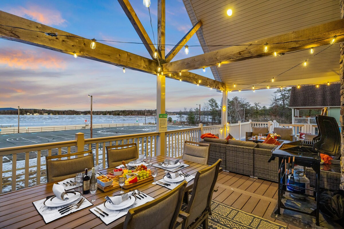 An outdoor deck features a dining table set for a meal, surrounded by chairs. Soft lighting from string lights enhances the space, with a view of the lake in the background. A barbecue grill and lounge seating complete the inviting atmosphere.