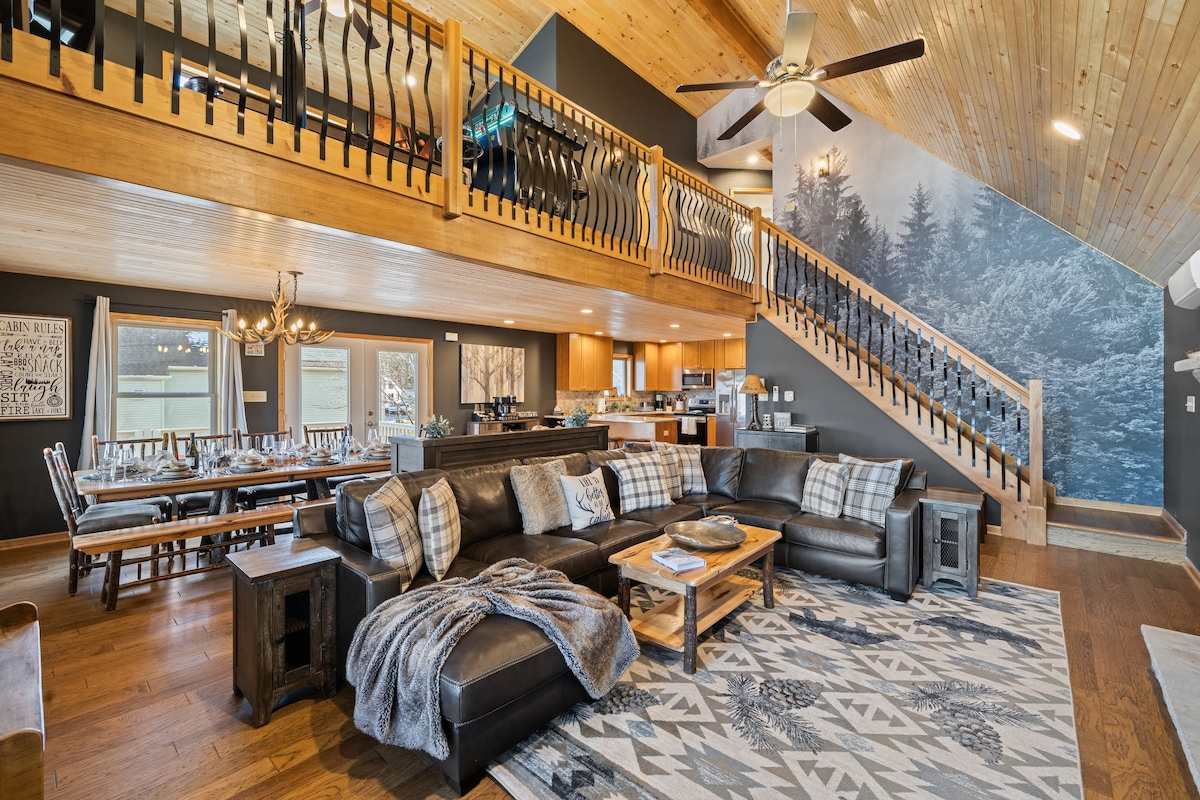 An open-concept living area features a spacious leather sectional sofa adorned with checkered pillows, centered around a wooden coffee table. Light wood accents and vaulted ceilings enhance the inviting atmosphere. A dining area is visible, with a long table set for gathering, while a staircase leads to the upper level.