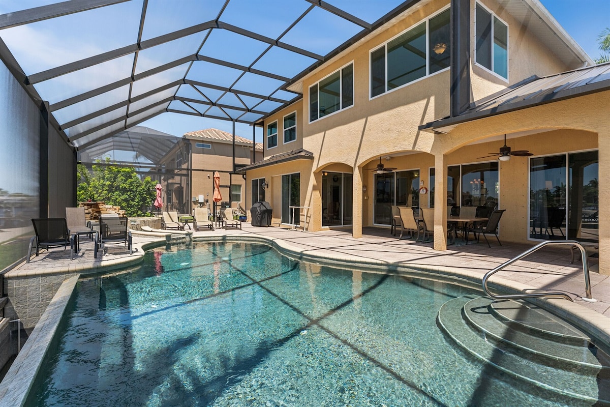 An expansive pool area is enclosed within a netted screen, showcasing a large swimming pool with a built-in spa. Multiple lounge chairs are positioned around the pool, while a covered patio offers shaded seating. The villa's large windows provide views of the outdoor space.