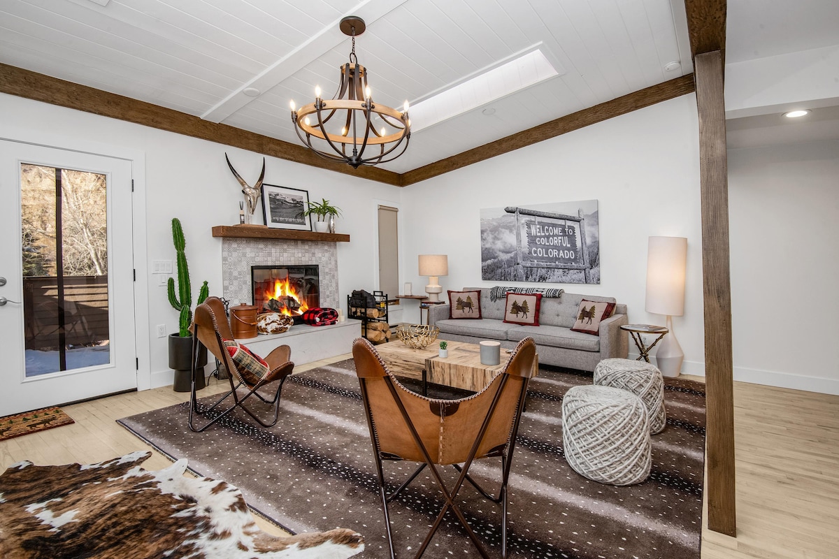 A welcoming living area features a comfortable sofa paired with armchairs, complemented by a rustic coffee table. A fireplace is central to the space, and large windows allow natural light to illuminate the room. Decorative elements include a cowhide rug and a modern chandelier.