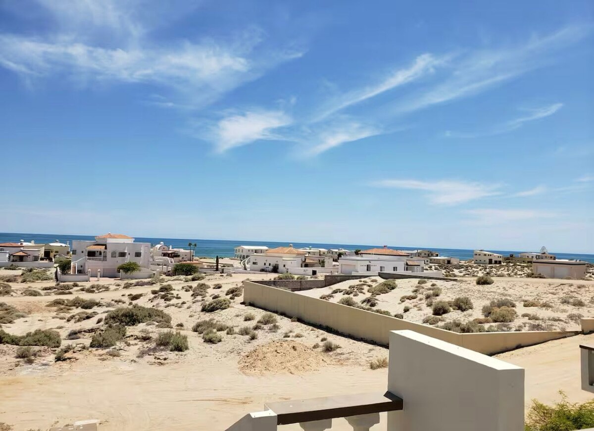 A panoramic view of the ocean and surrounding landscape is visible, with soft sandy terrain and a mix of residential buildings dotting the horizon. The clear blue sky features wispy clouds, enhancing the serene atmosphere.