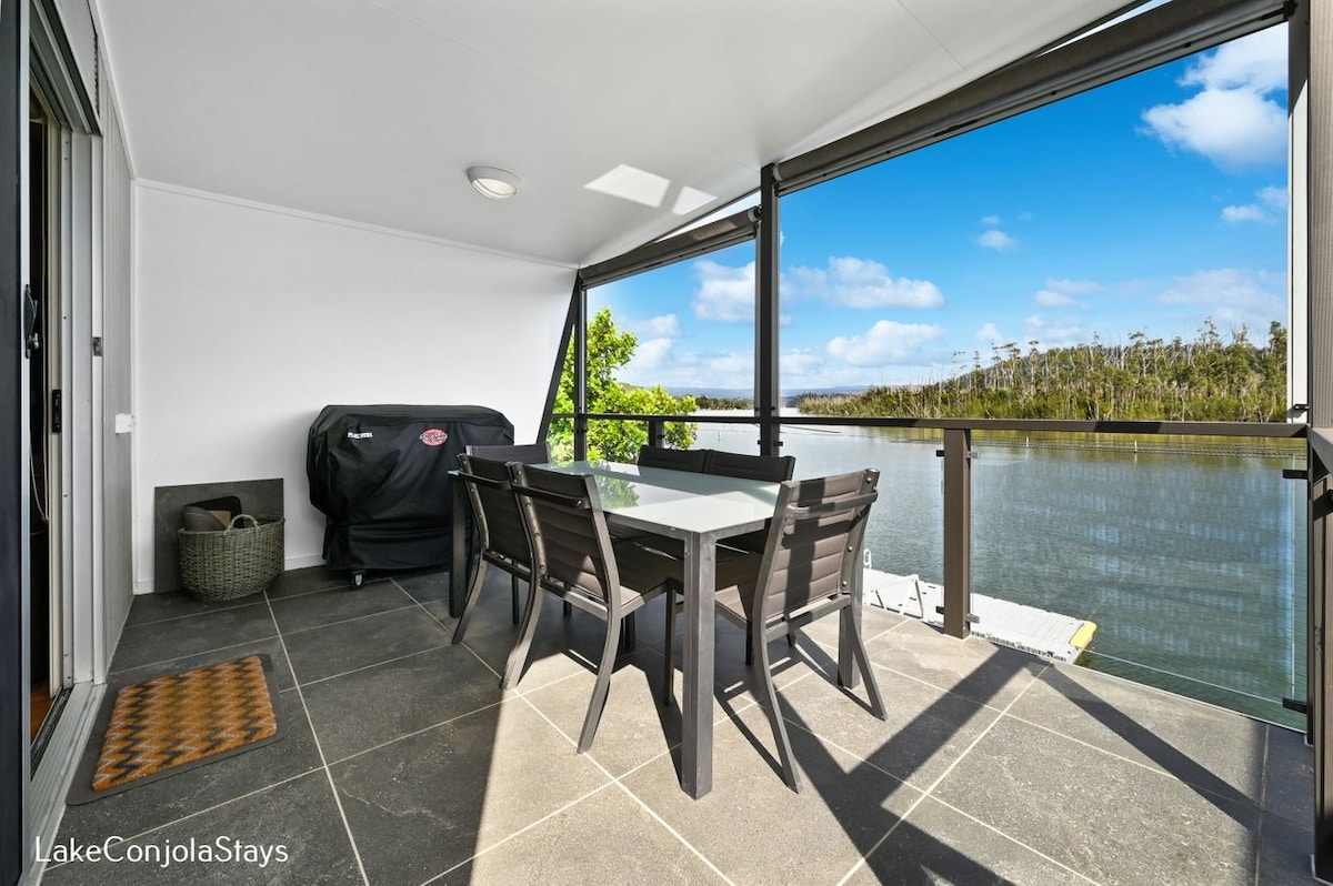 A covered balcony features a dining table with sleek black chairs, overlooking tranquil waters. A BBQ grill is stationed nearby, while large windows allow natural light to fill the space and offer scenic views of the surrounding nature.