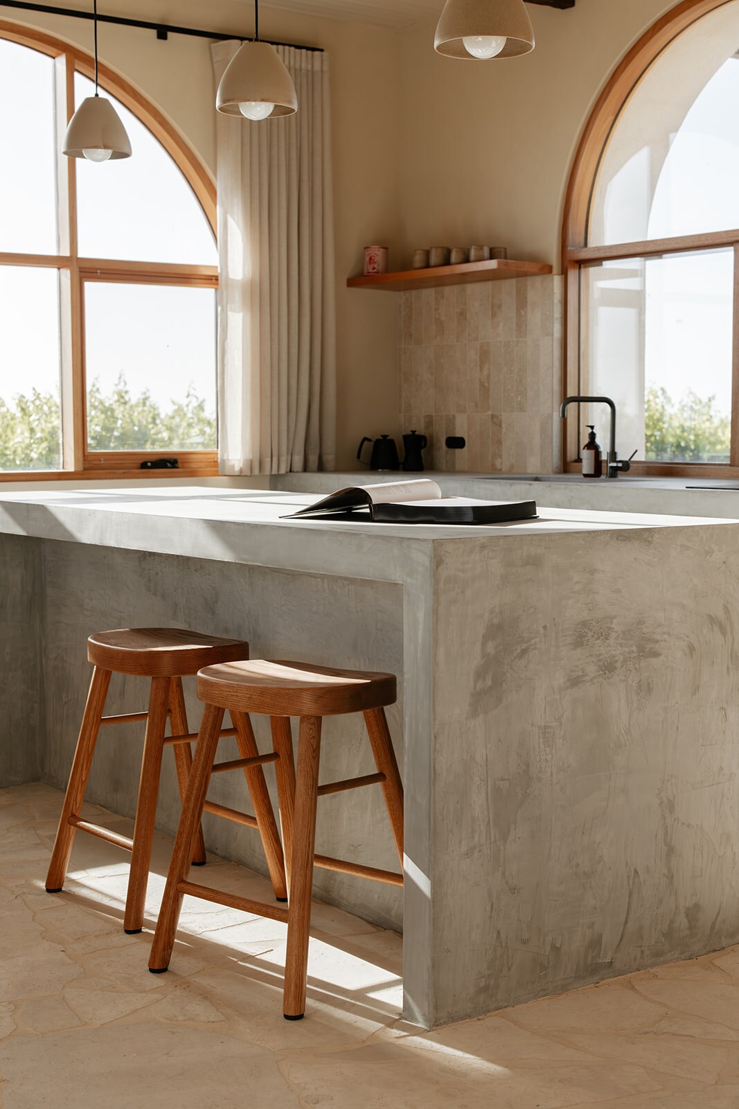 A modern kitchen features a concrete island with two wooden stools positioned beside it. Large arched windows allow natural light to fill the space, complemented by subtle pendant lighting hanging above.