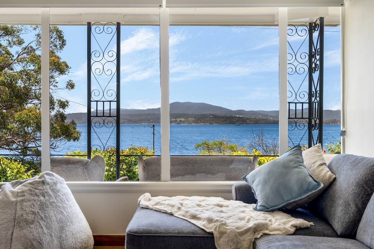 A bright living area offers a panoramic view of the tranquil Huon Valley waters through large windows. Plush seating, covered in soft textiles, complements the serene atmosphere, while greenery outside enhances the connection to nature.
