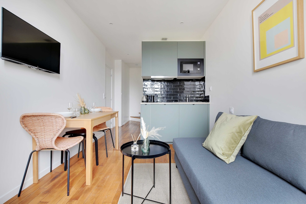 A modern living space features a comfortable sofa with a decorative pillow. A television is mounted on the wall across from a dining table set for two. The kitchen area contains sleek cabinetry, a microwave, and stylish backsplash tiles, contributing to the functional layout.