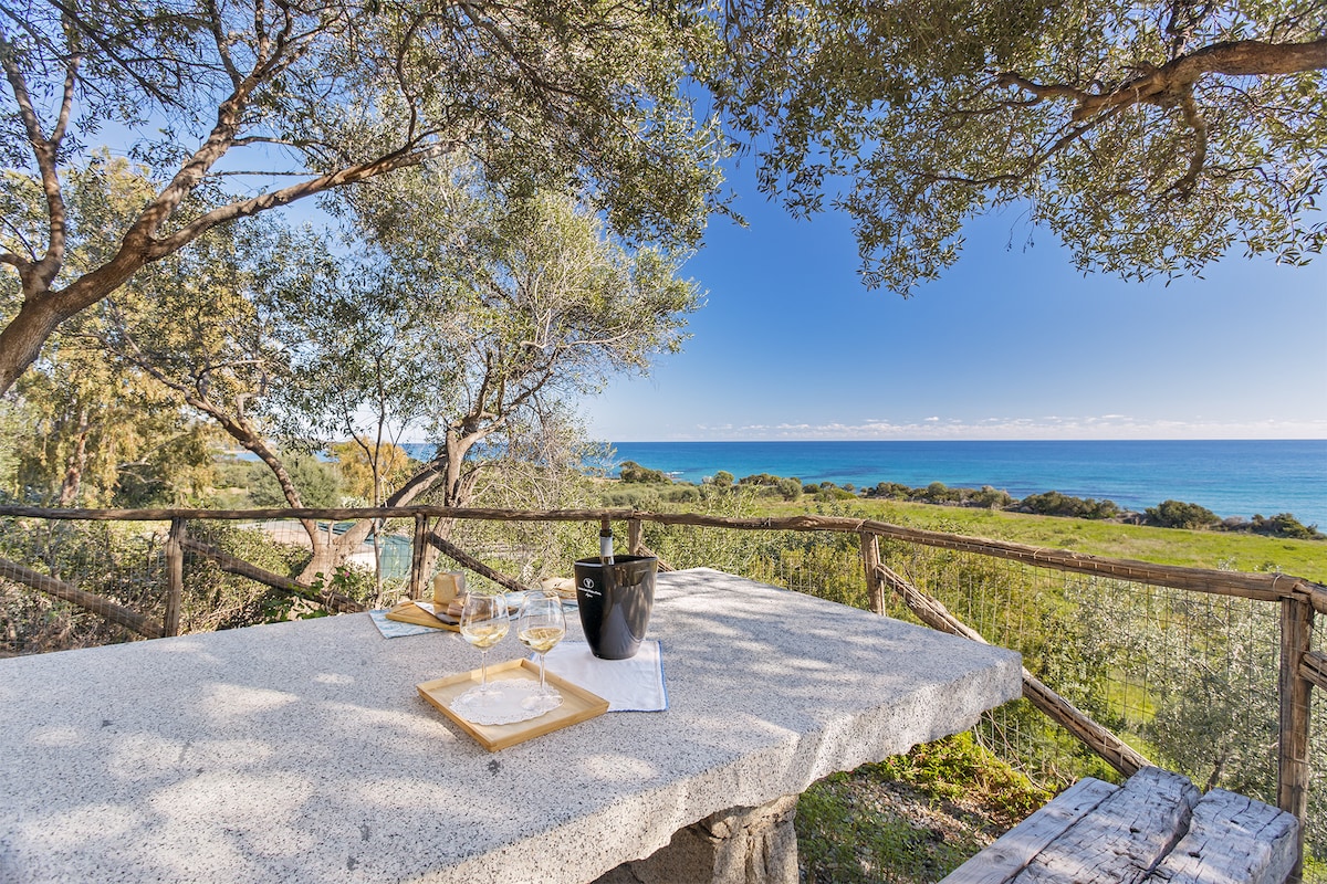 A stone table is set outdoors, overlooking the sea. Glasses and a bottle are arranged on a tray, while trees provide shade. The tranquil waters and lush greenery create a serene backdrop for relaxation.