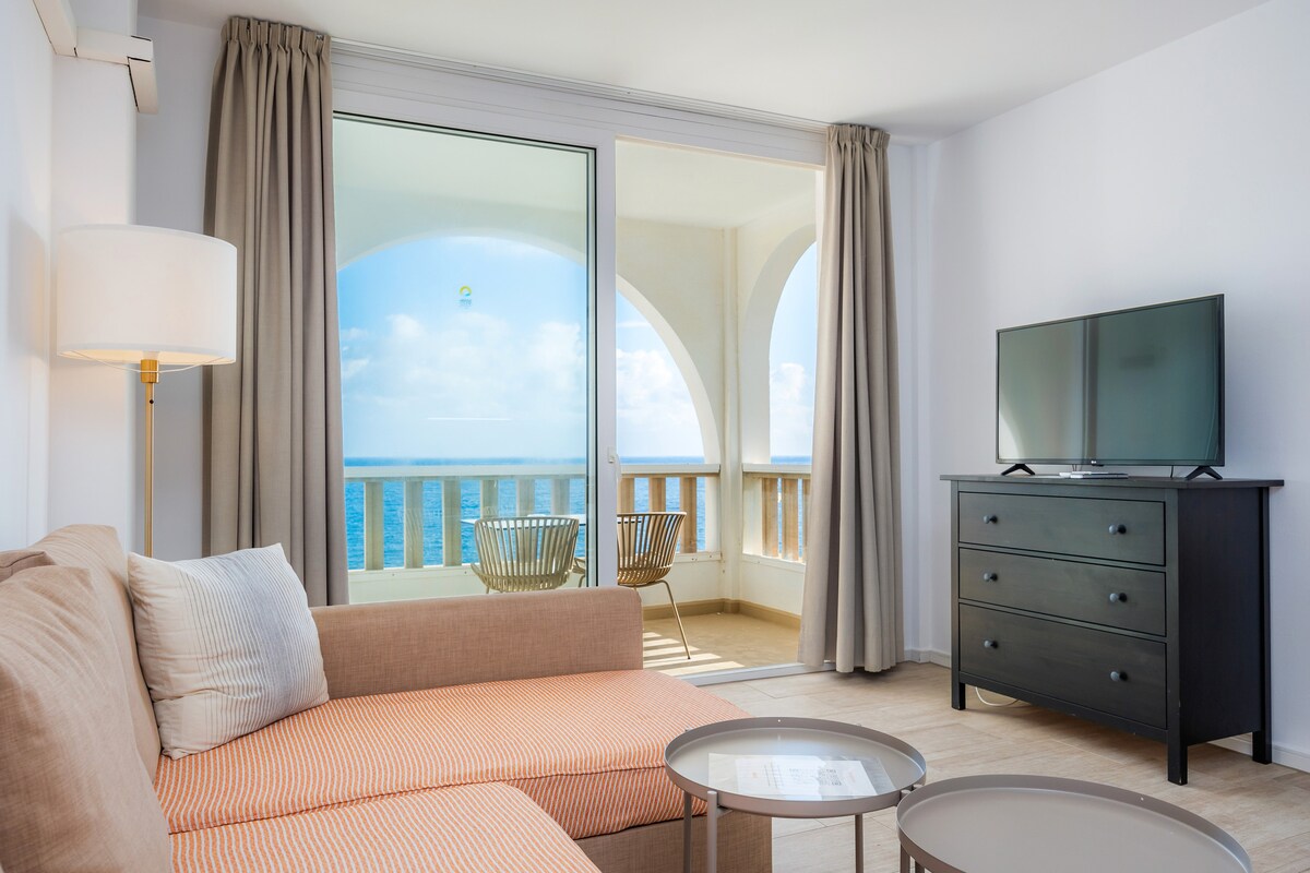 A bright living area features a comfortable sofa, a TV atop a dark dresser, and large glass doors leading to a private balcony. Outside seating on the balcony offers unobstructed views of the sea. Neutral colors and natural light create a refreshing ambiance.