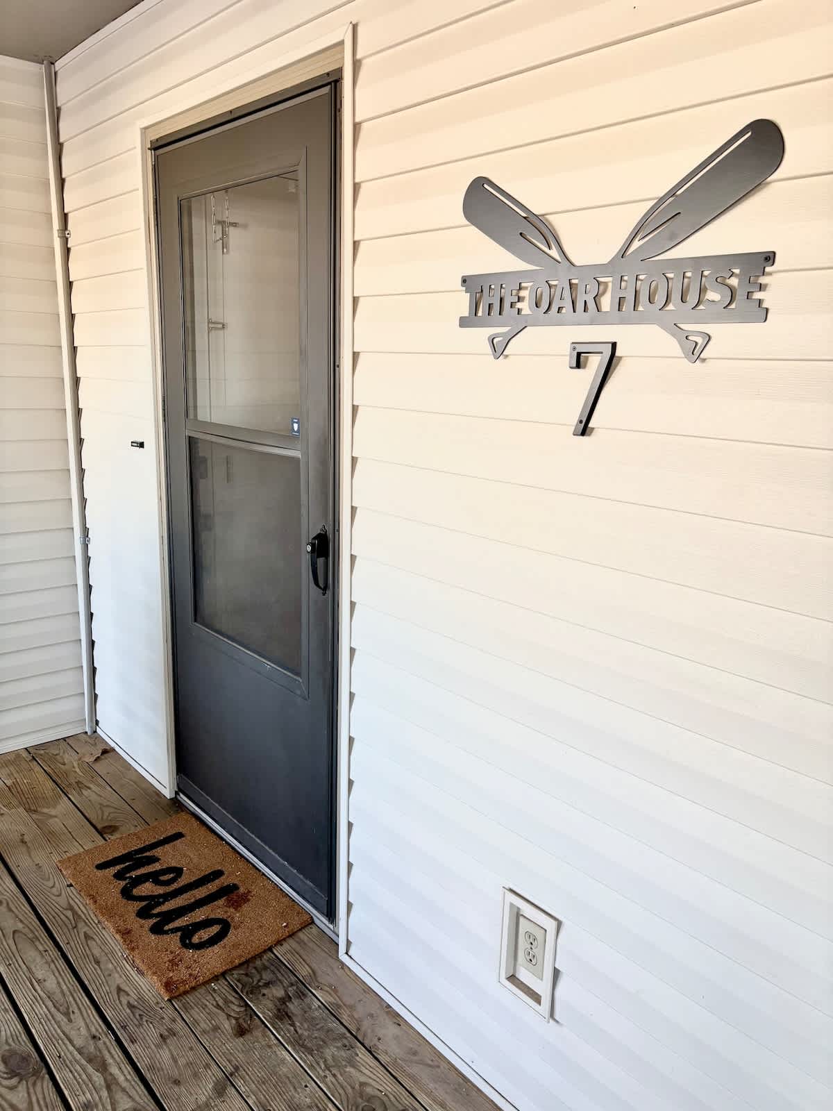 The entrance to the unit displays a sleek gray door beside a welcoming doormat that reads 'hello.' A stylish sign above features crossed oars, indicating 'The Oar House' and the number 7. The porch area is lined with hardwood flooring.