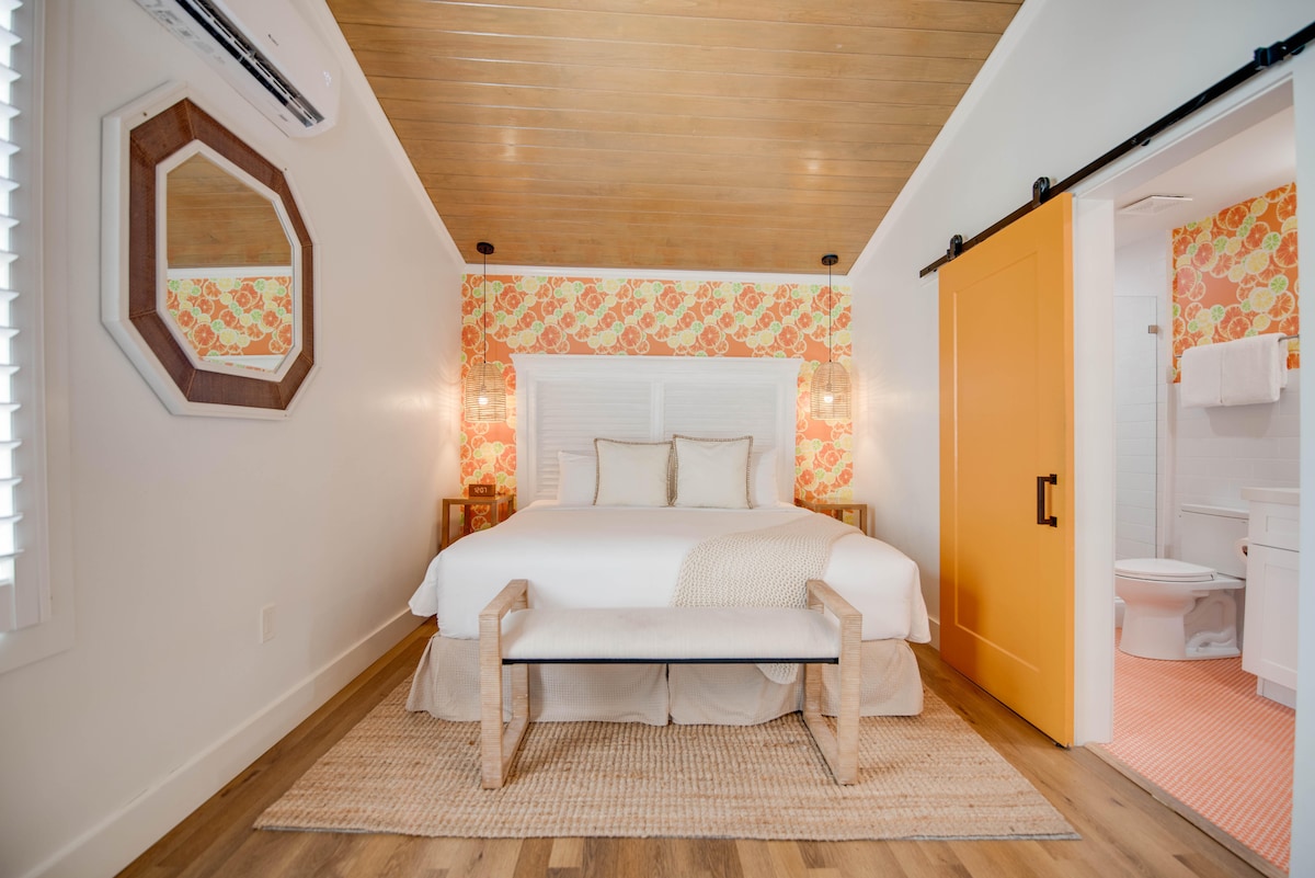 The studio features a bright sleeping area with a king-sized bed, adorned with light bedding. A natural wood bench sits at the foot of the bed, complemented by floral wallpaper and modern light fixtures. A sliding barn door leads to a stylized bathroom area.