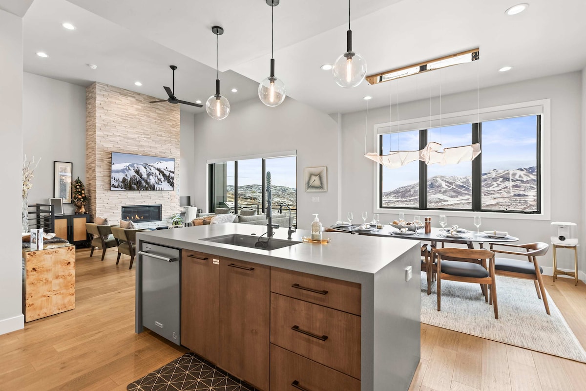 An open-concept kitchen and dining area feature modern appliances and sleek cabinetry. Large windows provide expansive views of the surrounding landscape. The dining table is elegantly set, with a cozy living space in the background, accented by a stone fireplace and contemporary lighting fixtures.