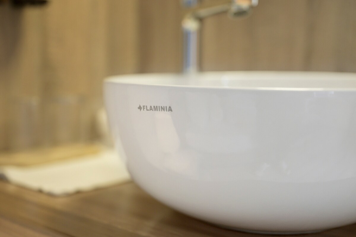 A contemporary washbasin with smooth, rounded edges is featured prominently. The sleek white ceramic contrasts against the warm wooden surface beneath. Minimalist glass cups are placed nearby, enhancing the clean aesthetic of the bathroom space.