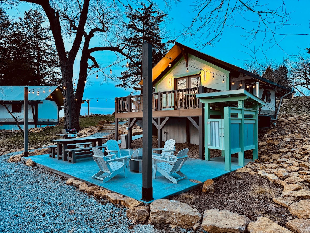 An inviting outdoor space features a large picnic table and several white adirondack chairs surrounding a fire pit. The backdrop includes a well-lit retreat structure with a covered balcony, complemented by twinkling string lights hanging above the patio area. Natural rock landscaping adds texture.