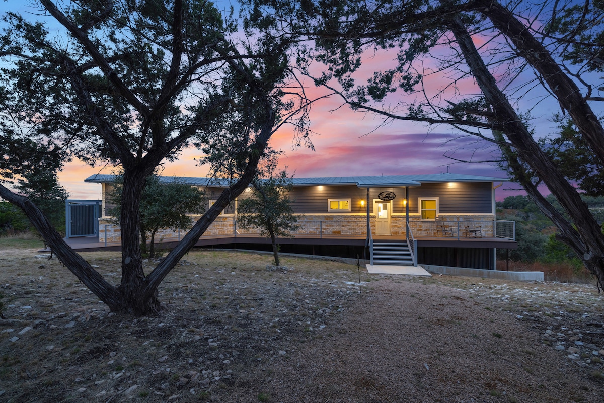 A two-level home is framed by trees under a vibrant sunset sky. An expansive deck features a railing and several outdoor chairs, inviting relaxation. Steps lead up to the entrance, and natural landscaping surrounds the property, highlighting its integration with the hill country.