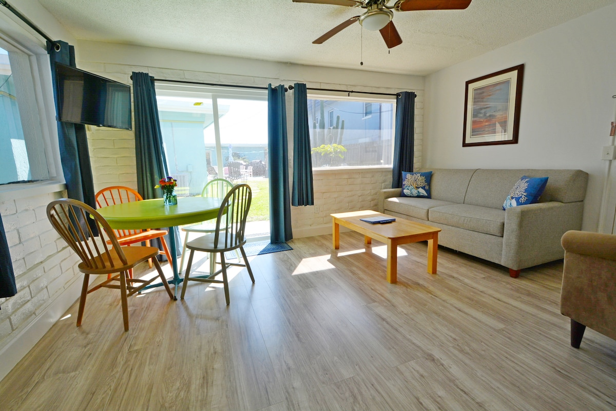 A bright living area features a comfortable sofa with decorative cushions, a wooden coffee table, and a dining set with vibrant chairs. Large windows allow natural light to fill the space, with a view of the outdoors framed by light blue curtains.