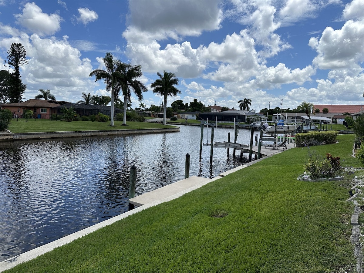 A serene waterfront scene showcasing calm waters bordered by lush green grass. Palm trees line the shore, providing a tropical atmosphere. A dock extends into the water, offering potential for boating activities, while fluffy clouds drift across the bright blue sky.