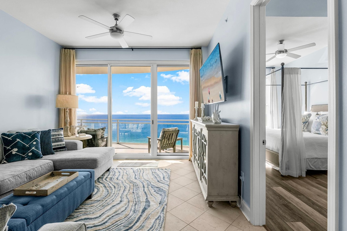 A bright living area is highlighted by large windows with stunning ocean views. A comfortable sectional couch faces a wall-mounted television, while light blue walls complement the coastal decor. A cozy side table and lamp provide sufficient lighting, contributing to a relaxed atmosphere.