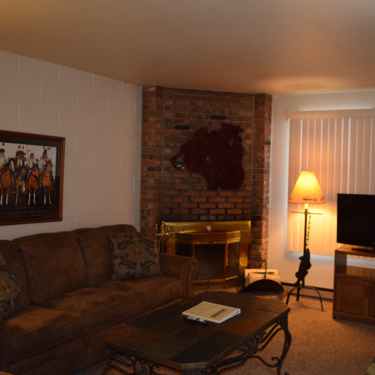 A living room features a brown sofa adorned with decorative pillows, positioned near a brick fireplace. A side lamp provides warm lighting, and a TV is situated on a wooden stand. A coffee table is centered, accompanied by a storage unit in the background.