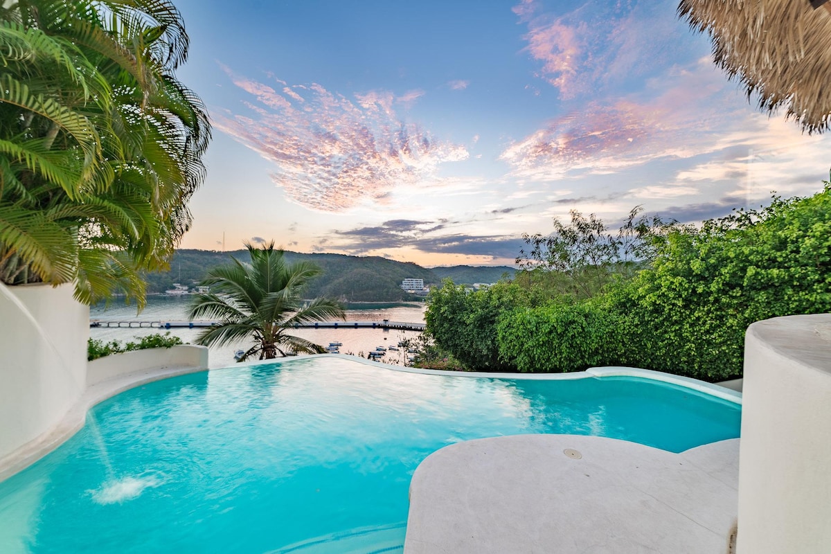 A tranquil infinity pool is set against a backdrop of lush greenery and palm trees. The pool water reflects the vibrant colors of the sunset sky, with gentle ripples creating a serene ambiance. Panoramic ocean views are visible in the distance.