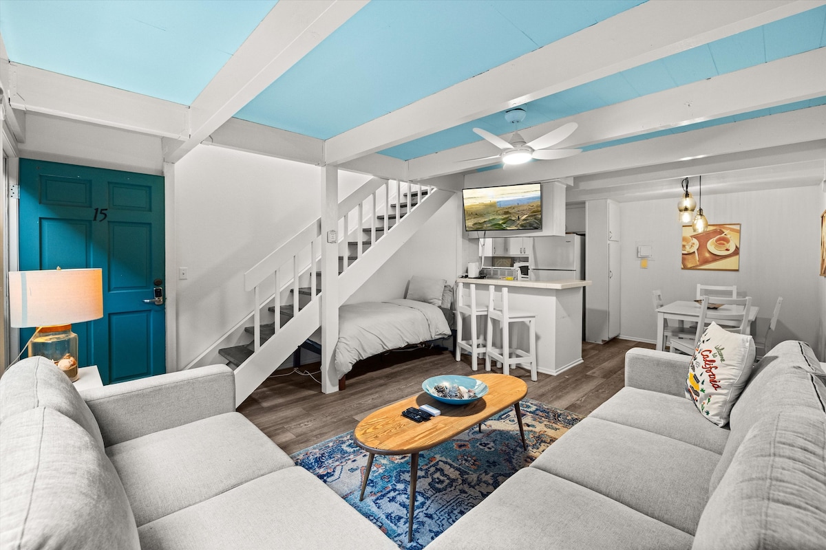 The open living area features a light grey sectional sofa facing a wooden coffee table. A ceiling fan gently rotates above, while a flat-screen TV is mounted on the wall. The turquoise ceiling complements the blue accent door, inviting a fresh and airy feel.