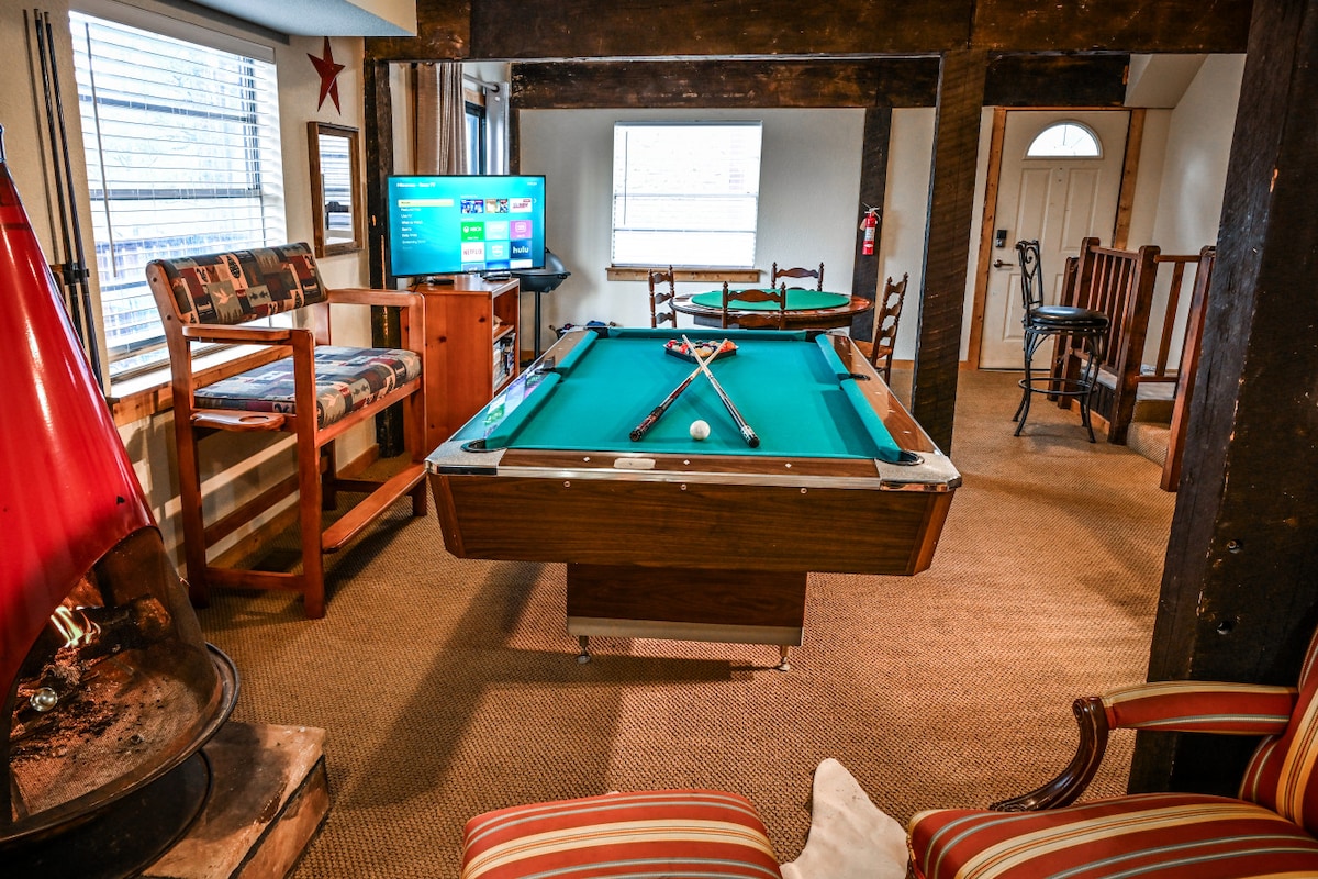 A game room is captured, featuring a billiards table at its center. A television is positioned nearby, while a cozy seating area consists of a bench and chairs. Natural light enters through a window, creating a welcoming environment with a visible fireplace in the corner.