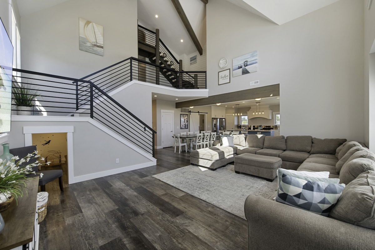 A spacious living area is presented, featuring a large sectional sofa arranged around a soft area rug. Natural light fills the room through expansive windows, while a contemporary staircase leads to the upper level. Open sight lines extend into the dining and kitchen areas.