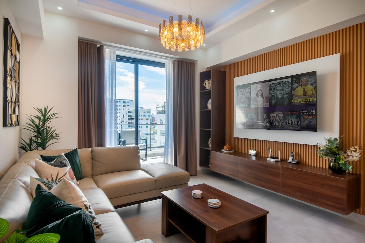 A modern living room is showcased, featuring a spacious sofa adorned with decorative cushions. A wall-mounted Smart TV is displayed alongside wooden shelving. Natural light filters through large glass doors that lead to a balcony, while a chandelier hangs from the ceiling, enhancing the elegant decor.