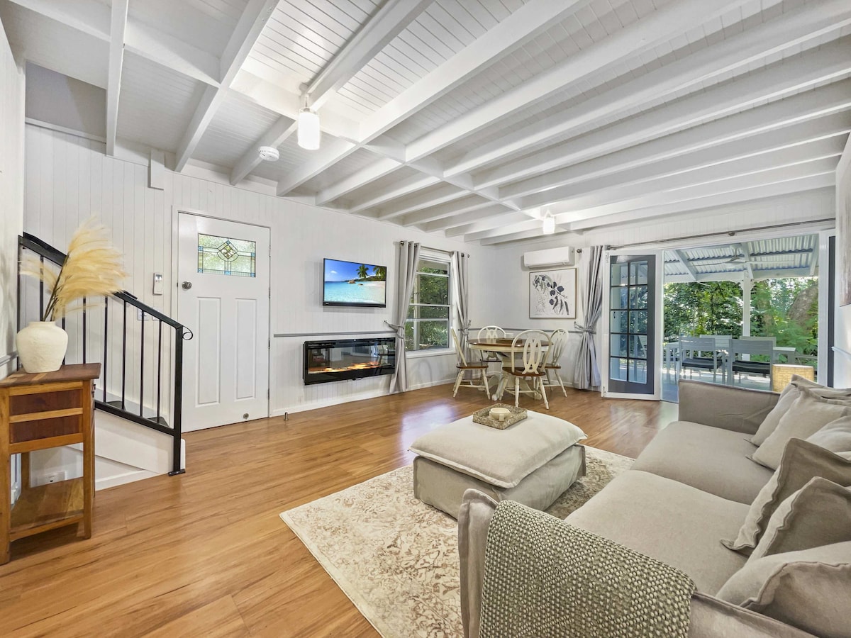 An open-plan living area features exposed timber ceilings and a cozy lounge with a large sectional sofa. Natural light floods the space through large windows. A dining area is set for four, while a flat-screen TV and an electric fireplace add to the comfort.