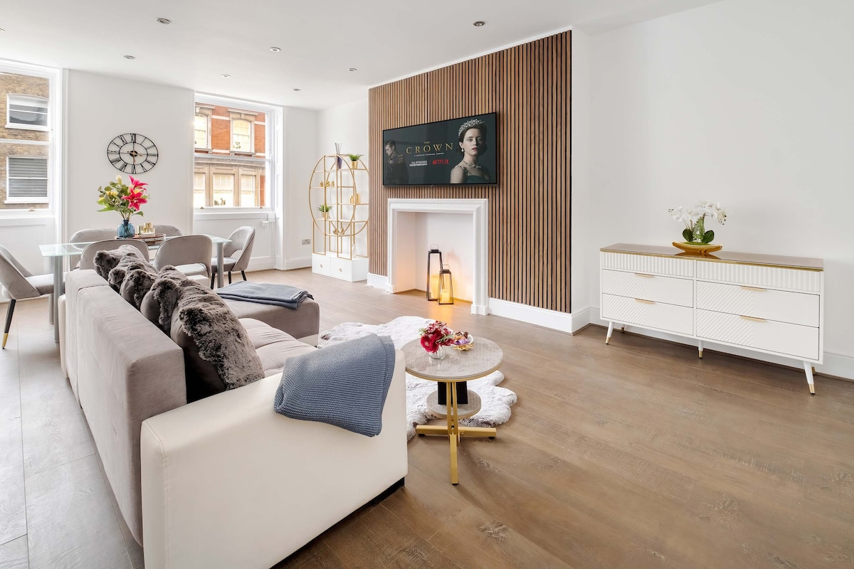 An open-concept living area features a combination of light hardwood flooring and contemporary furniture. A sofa with plush cushions faces a large flat-screen TV mounted on a feature wall. A stylish coffee table sits atop a soft area rug, with decorative elements enhancing the space.