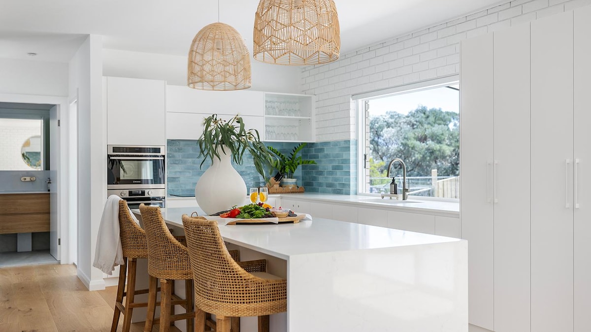 A modern kitchen features white cabinetry and a large island with natural wood seating. Two woven pendant lights hang above the island. A window provides a view of greenery, and a vase with a plant adds a touch of nature to the space.