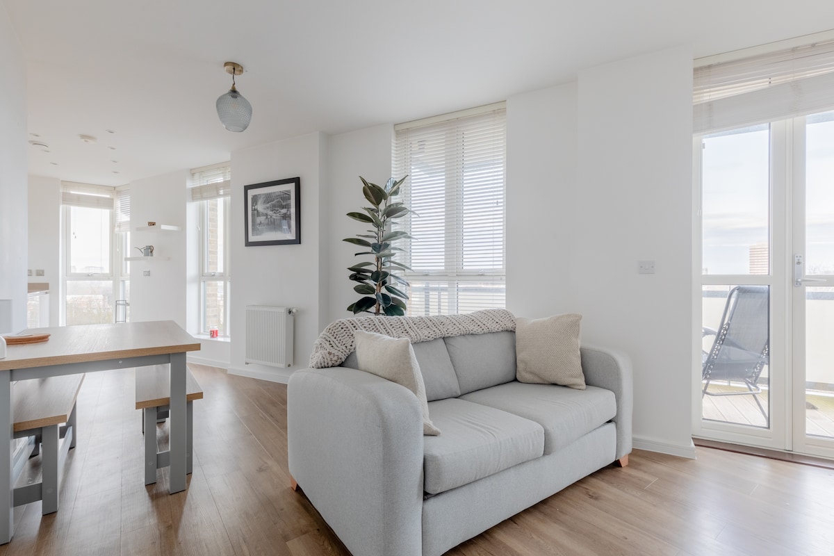A combined living and dining area is presented with large windows, allowing natural light to fill the space. A light grey sofa is adorned with soft cushions, while a simple dining table is visible nearby. A potted plant adds a touch of greenery to the room.
