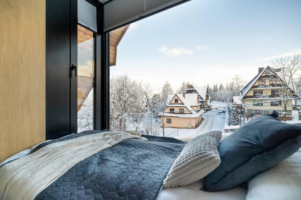 A cozy bedroom features a spacious bed adorned with soft bedding and decorative pillows. A large window provides a view of snow-covered trees and houses, highlighting the serene winter landscape. Natural light fills the room, enhancing its warm and inviting ambiance.