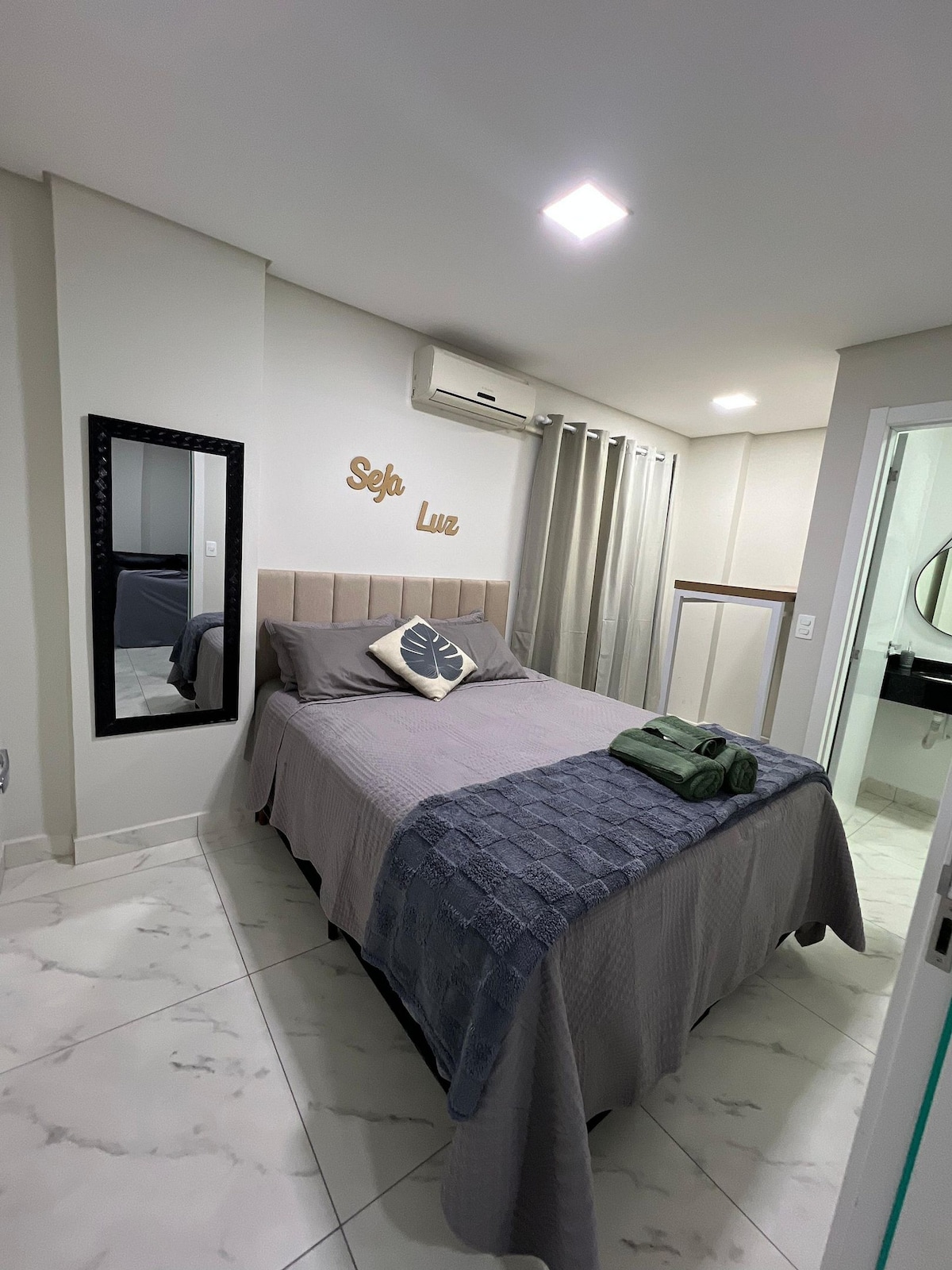 A cozy bedroom features a spacious bed with soft bedding and a gray throw. A mirror adorns one wall, reflecting the modern décor. Natural light enters through curtain-covered windows, complementing the neutral color palette. A small decorative sign is displayed above the bed.
